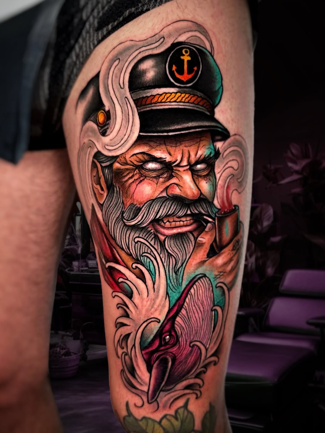 Colorful sailor captain portrait tattoo on thigh featuring a bearded man in naval hat with anchor emblem, ornamental scrollwork, and coral, green, and red tones.
