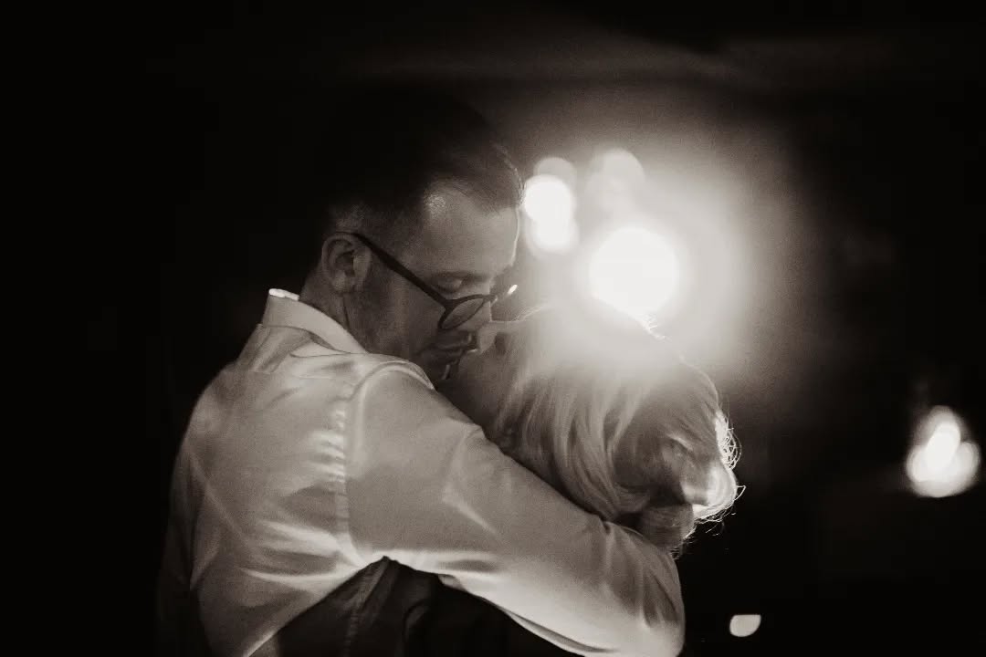 Man with glasses holding a dog against his chest in monochrome lighting, showcasing a tattoo on his forearm.