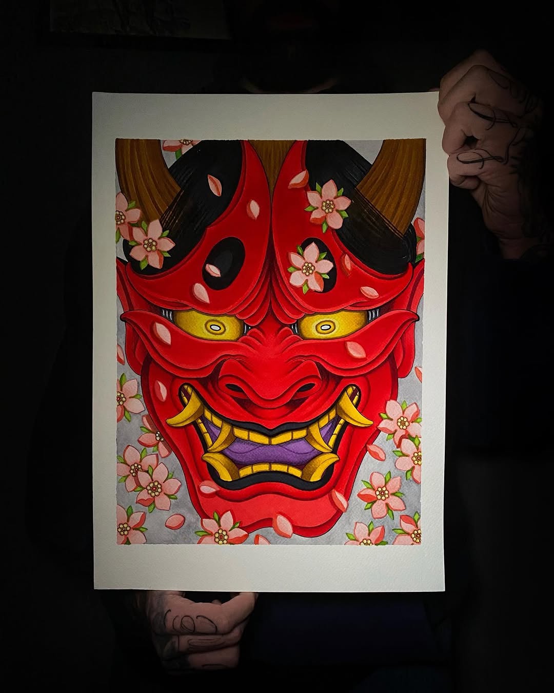 Red demonic mask with yellow eyes and gold fangs surrounded by pink flowers and brown hair, displayed on white paper.