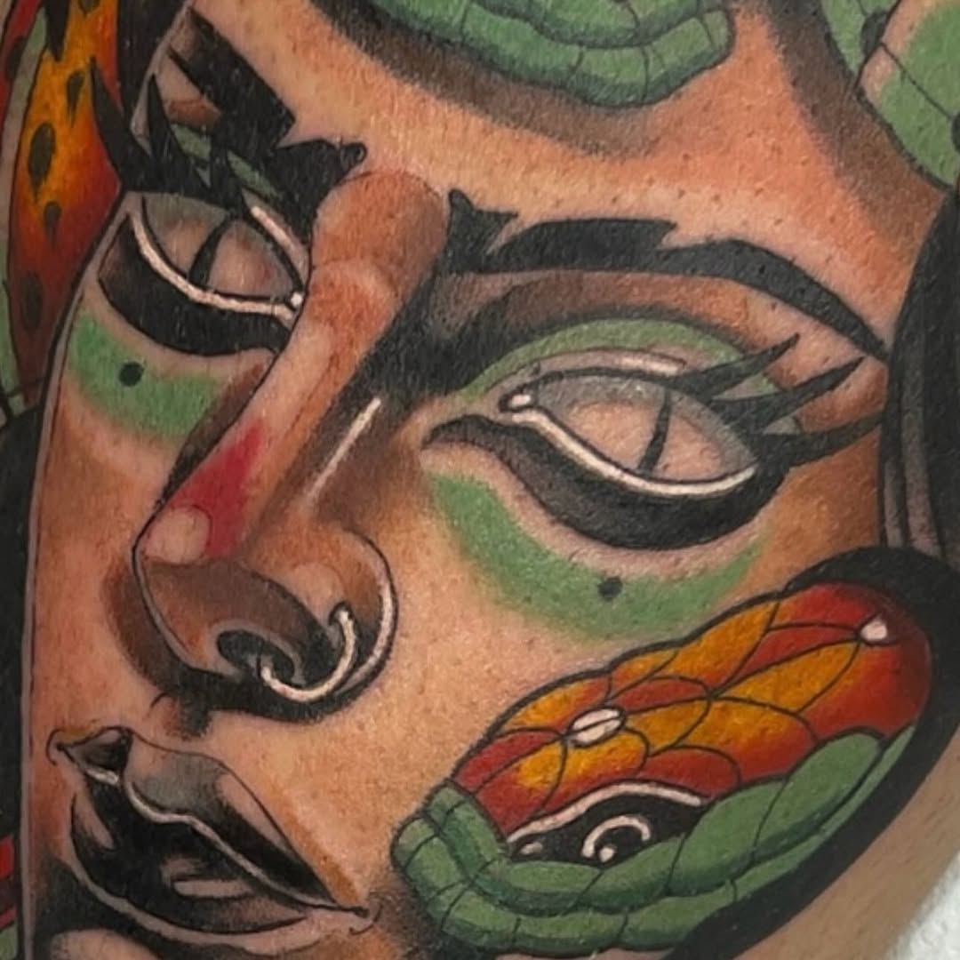Colorful facial tattoo featuring green and red leaf designs, black eyeliner details, and orange-red butterfly motifs on the cheek and forehead area.