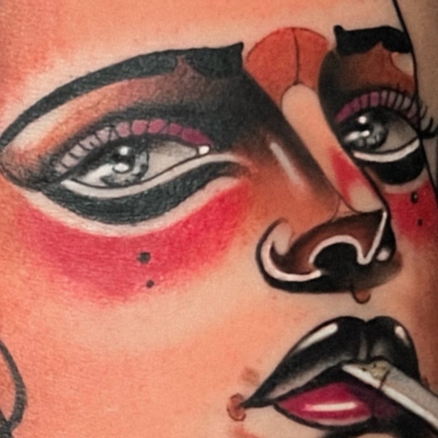 Colorful face tattoo featuring dramatic eye makeup with black and red eyeshadow, stylized nose, and bold black lips with white highlight on upper arm.