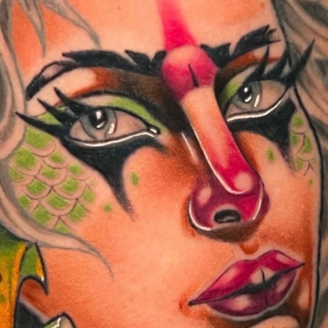 Colorful portrait tattoo on arm featuring a woman's face with green eyeshadow, magenta nose and lips, scale patterns, and orange skin tones.