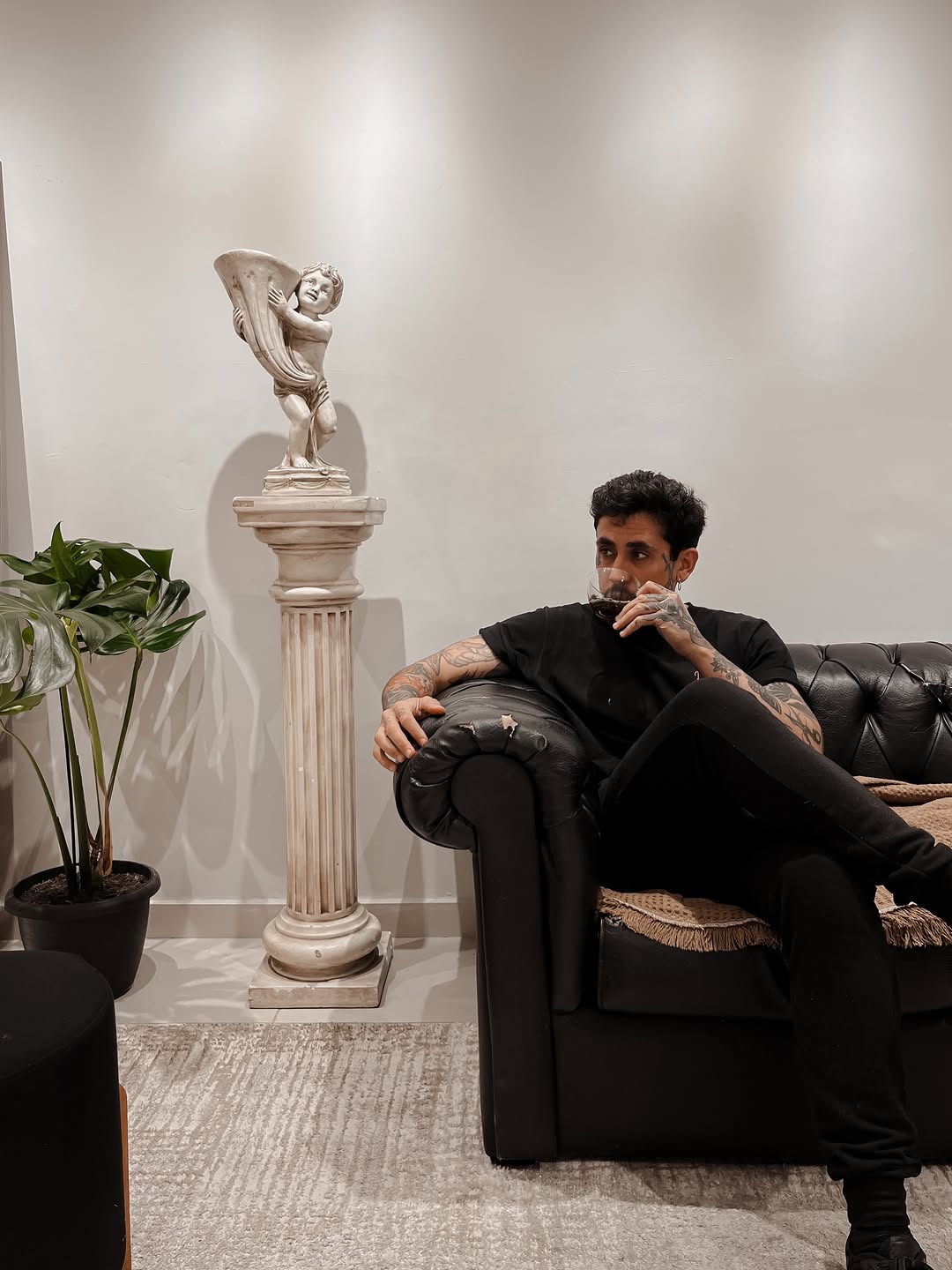 Man wearing black clothing seated on leather couch with visible arm tattoos next to decorative pedestal and potted plant in modern interior space.