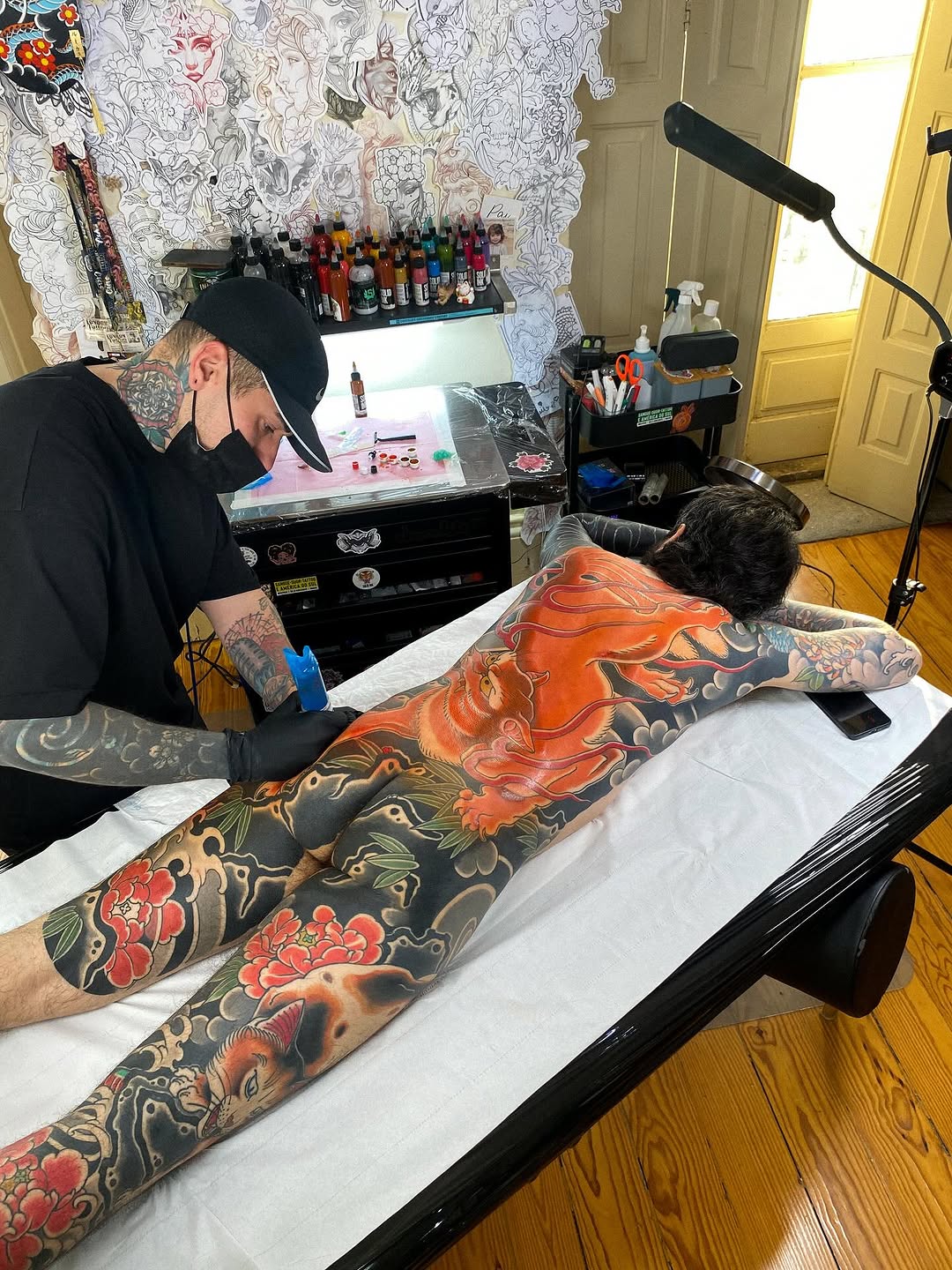 Tattoo artist working on client's back and legs featuring large red and orange dragon with floral designs in vibrant colors.