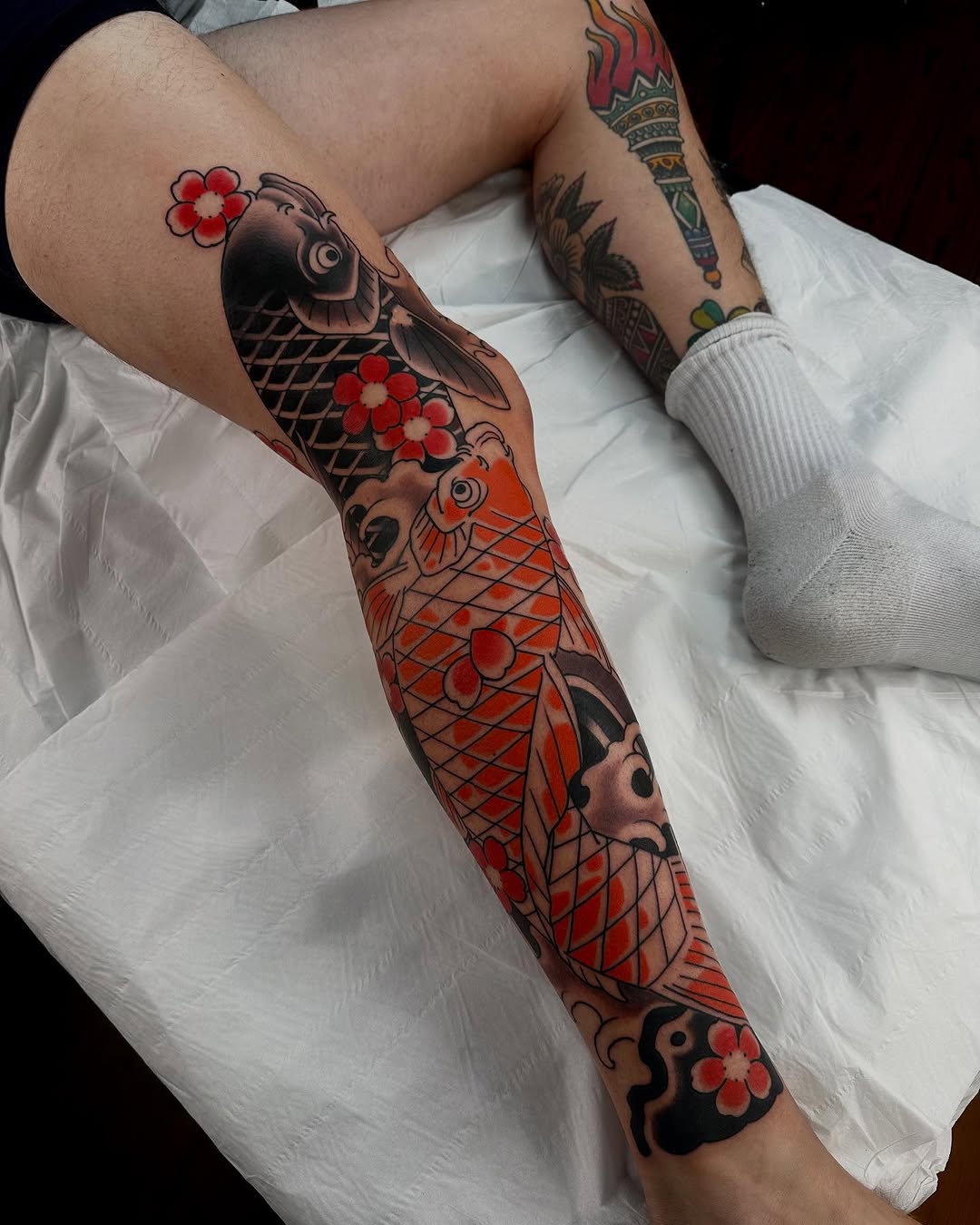 Full-leg sleeve tattoo featuring three koi fish in red and black with geometric scales, red flowers, and dark ornamental elements on the thigh and calf.