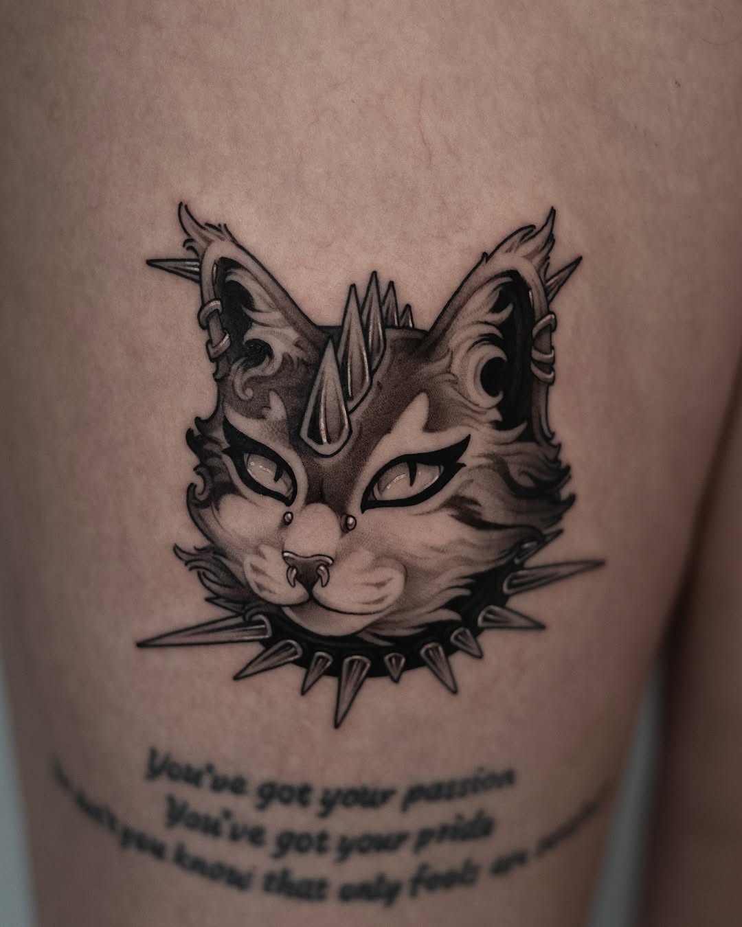 Decorative cat face with pointed ears, mystical markings, spiked collar, and text below on upper arm in black ink.