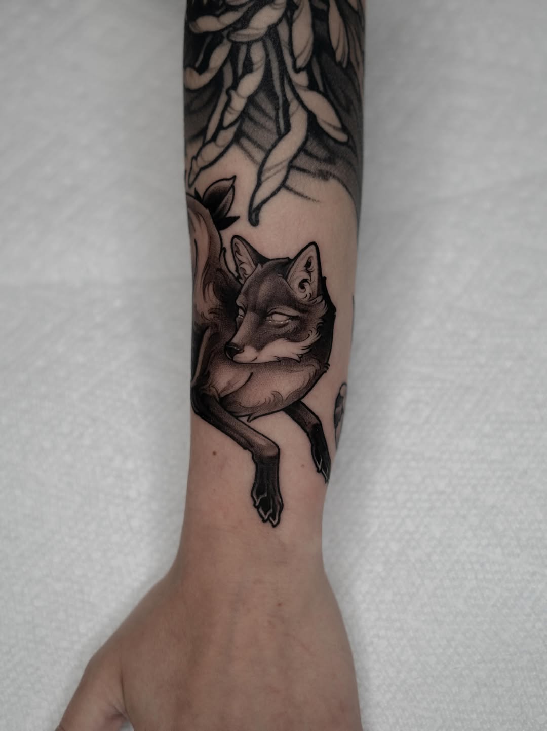 Tattoo on a forearm featuring a fox with closed eyes and a fluffy tail, surrounded by flowing botanical elements in black and brown ink.