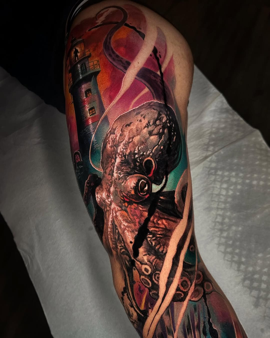 Colorful sleeve tattoo featuring an octopus with glowing eyes, a lighthouse, swirling ribbons, and decorative patterns in red, orange, teal, and purple tones.