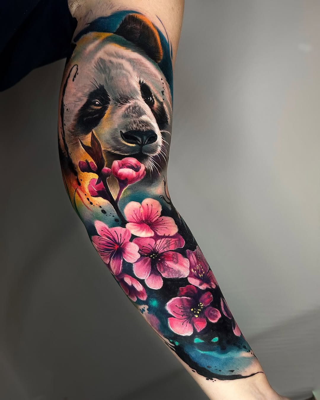 Colorful panda head tattoo on forearm with pink flowers, blue and gold accents, and splatter effects covering the upper arm to wrist.