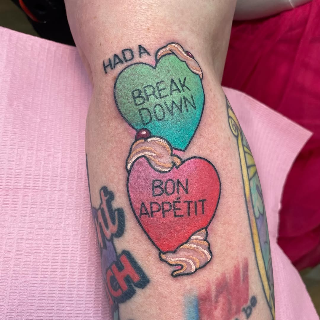 Two cupped hands holding a turquoise heart labeled "HAOA BREAKDOWN" and a red heart labeled "BON APPÉTIT" tattooed on a leg.