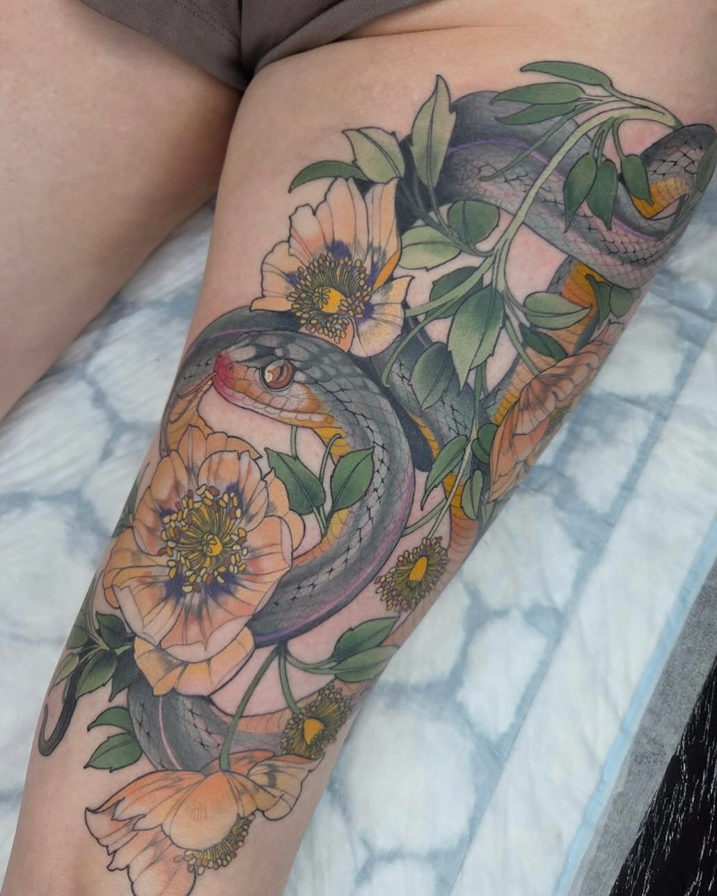 Colorful thigh tattoo featuring two snakes intertwined with peach and white flowers, green leaves, and yellow centers on skin.