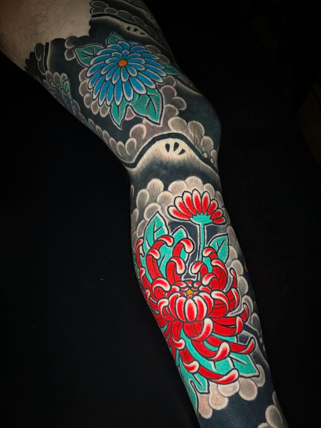 Colorful floral sleeve tattoo featuring blue and red flowers with green leaves on black background, wrapped around the arm with decorative cloud motifs.