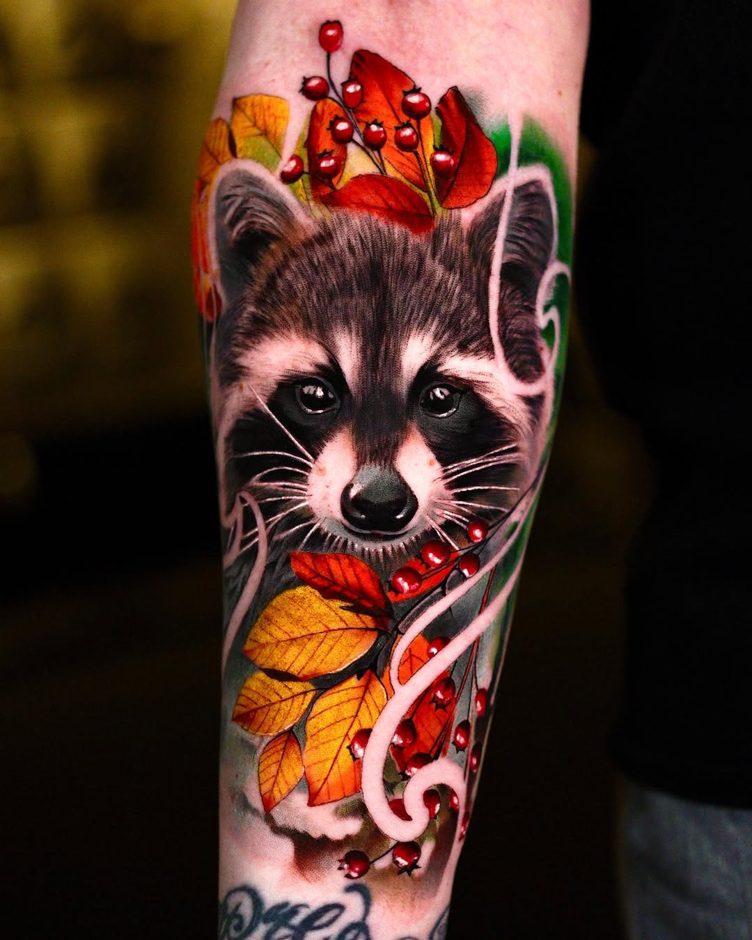 Raccoon head tattoo with autumn leaves and red berries in orange and red tones on the forearm.