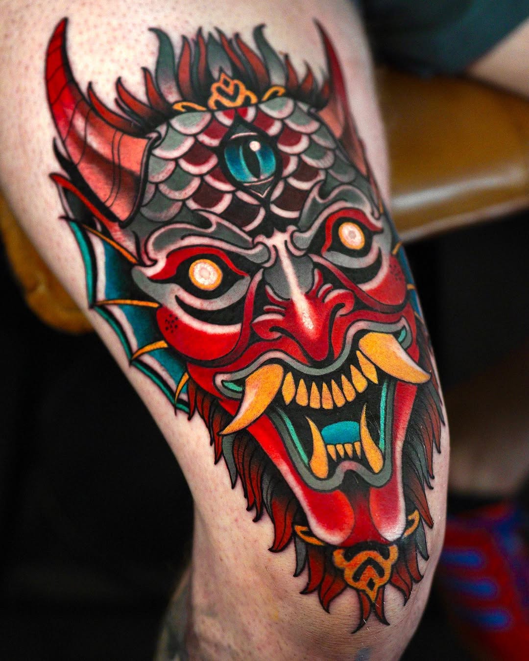 Colorful demon head tattoo on upper arm featuring red face, horned crown, scaled forehead, multiple eyes, and orange fangs with teal and yellow accents.
