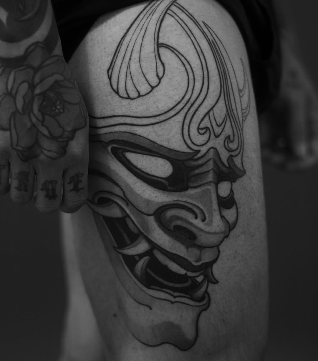 Theatrical mask tattoo on upper thigh featuring dramatic facial features with ornate curved lines, rendered in black ink on pale skin.