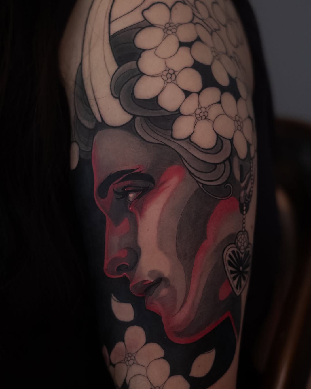Profile of a red-faced figure with decorative flowers and ornamental elements in black and white, tattooed on the upper arm.