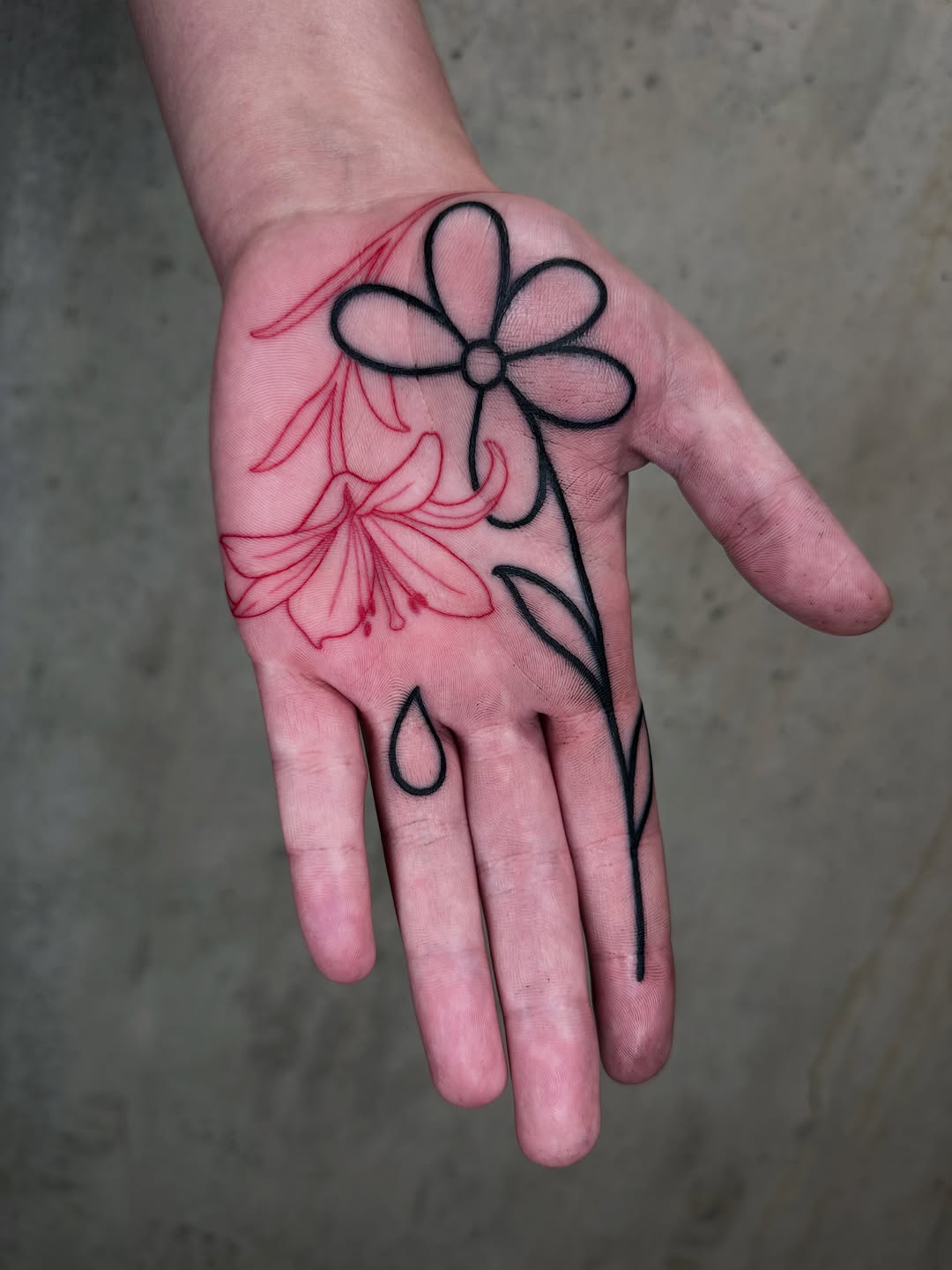 Hand tattoo featuring a black-outlined daisy flower and a red detailed flower with flowing petals and stems on the palm and back of hand.