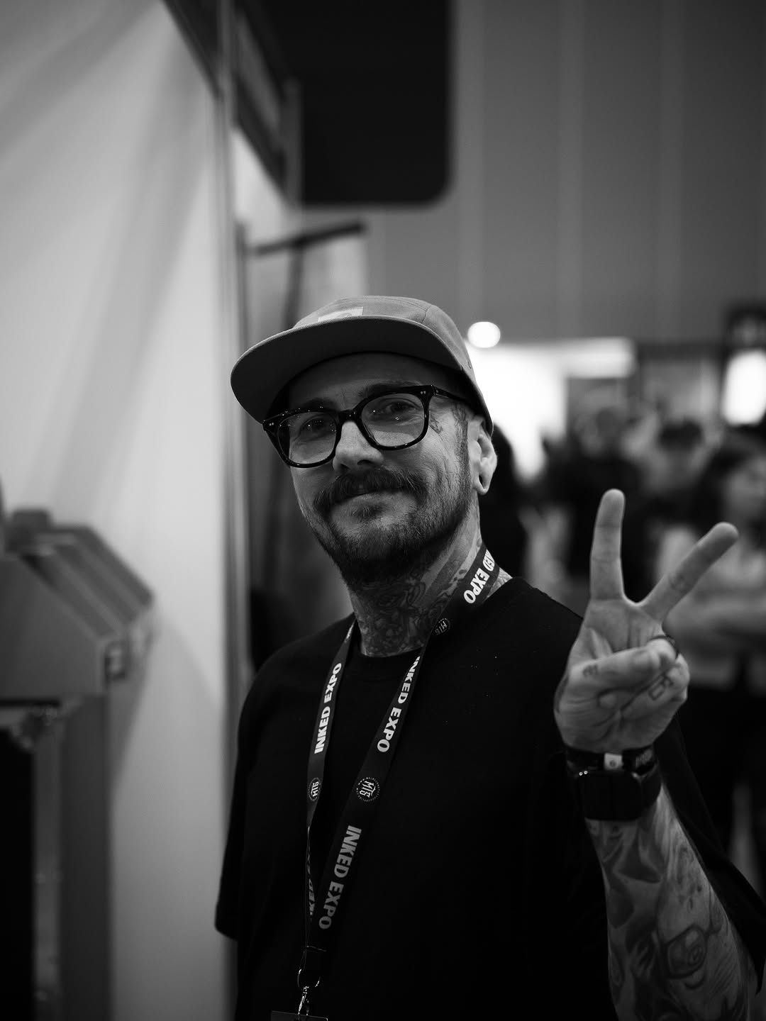 Bearded man wearing glasses and a baseball cap displays an arm tattoo while making a peace sign at what appears to be a professional event.