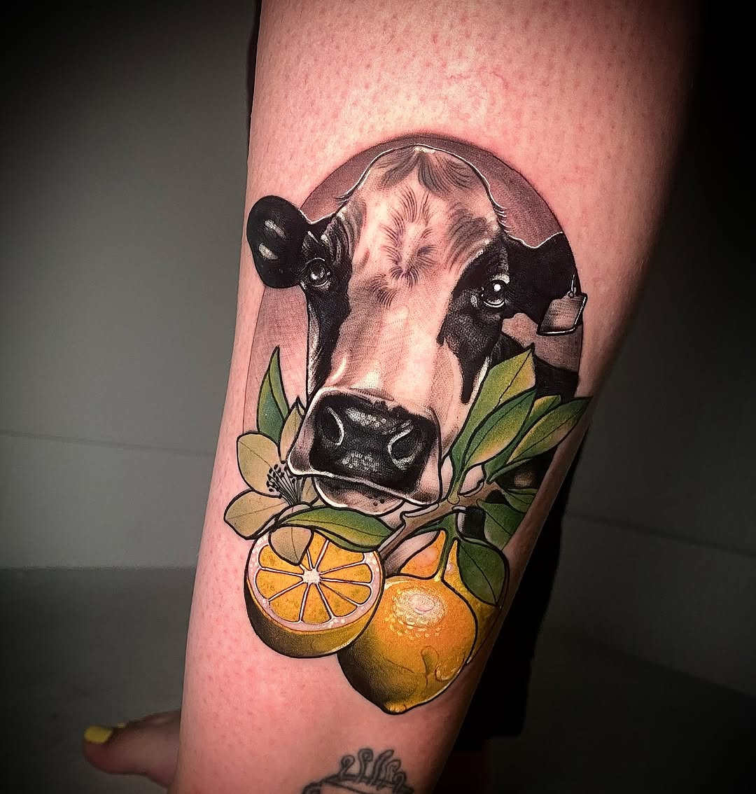 Cow's head with black and white shading surrounded by orange citrus fruits and green leaves, tattooed on the upper leg.