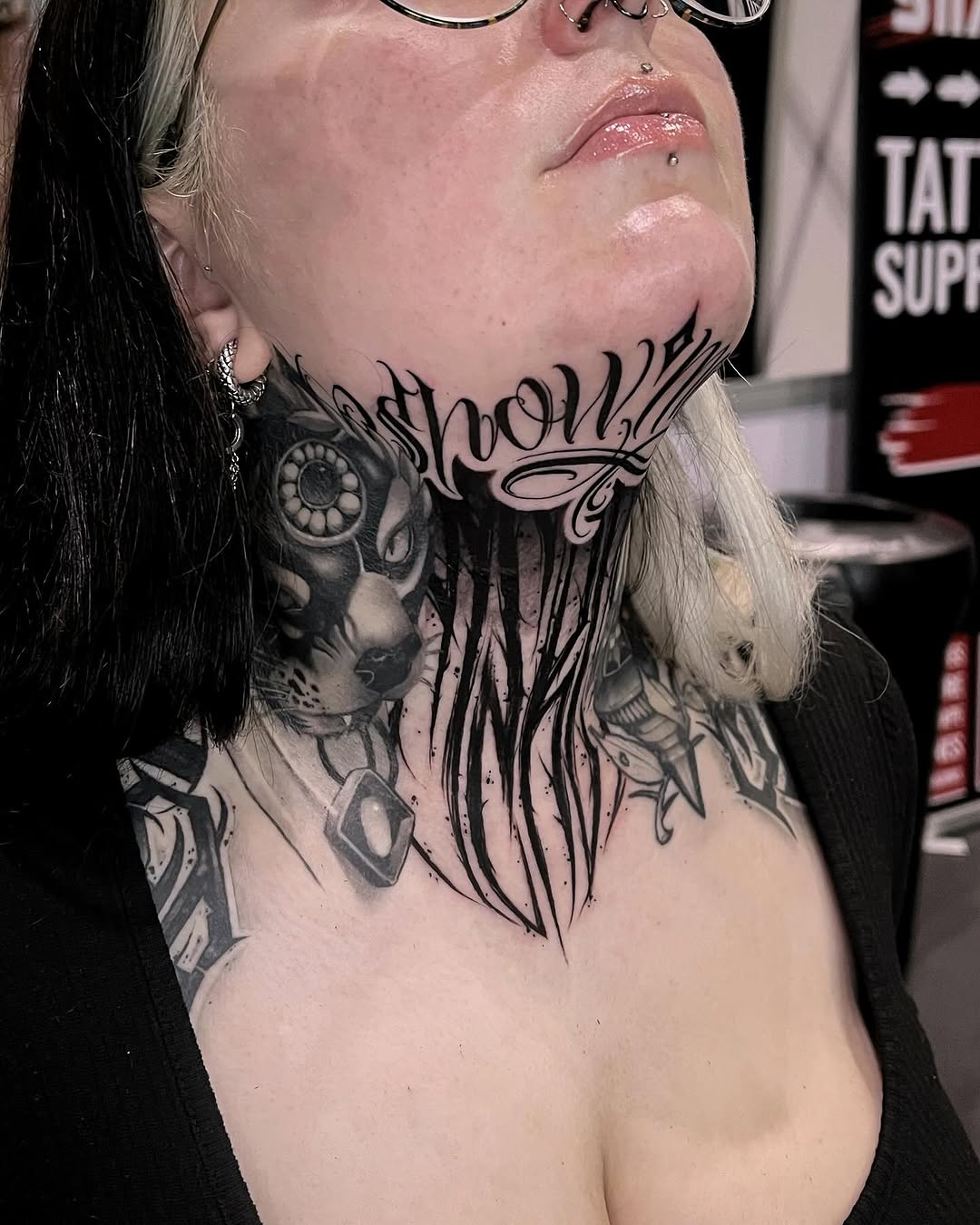 Large-scale neck and chest tattoo featuring an eye, flowing script text, and ornamental designs in black ink on pale skin.