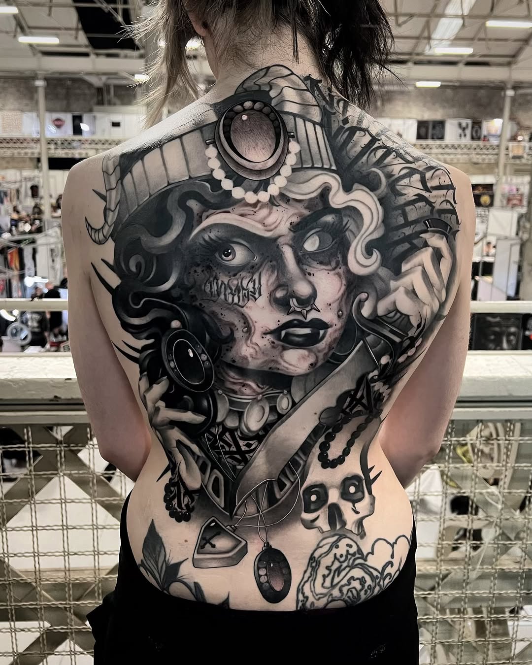 Large back tattoo featuring an ornate female face with decorative elements, pearls, and smaller symbolic imagery below in black and gray ink.