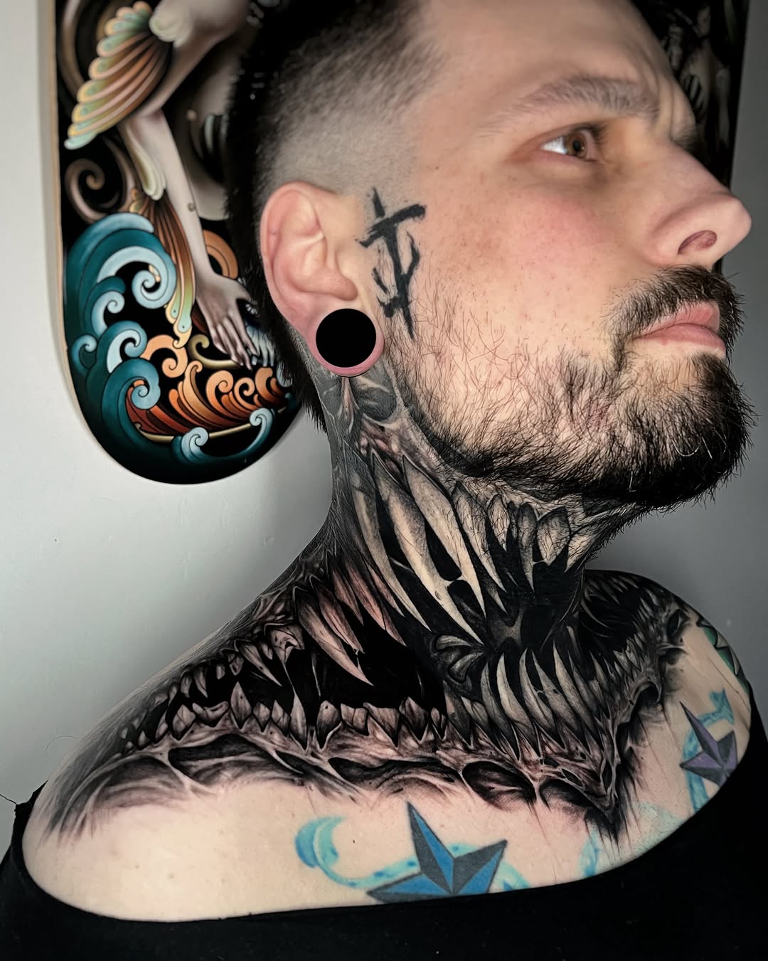 Man with large feathered neck tattoo in black and grey, ear gauge, facial tattoo, and colored chest and arm tattoos displayed in profile.