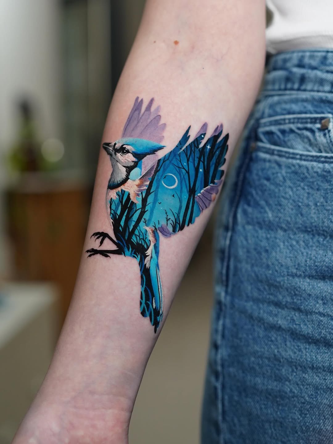 Blue jay tattoo with purple watercolor wings and black line details on a forearm, featuring the bird perched with detailed head and extended tail feathers.