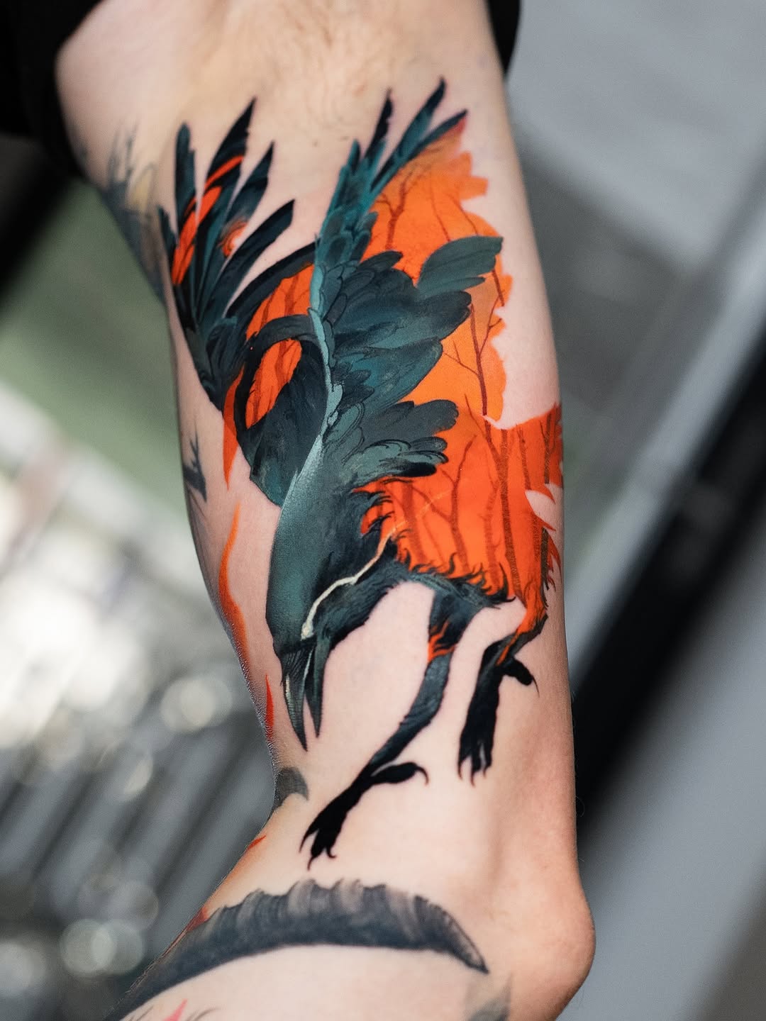 A colorful bird tattoo with dark teal and black plumage and bright orange-red wings on a person's forearm.