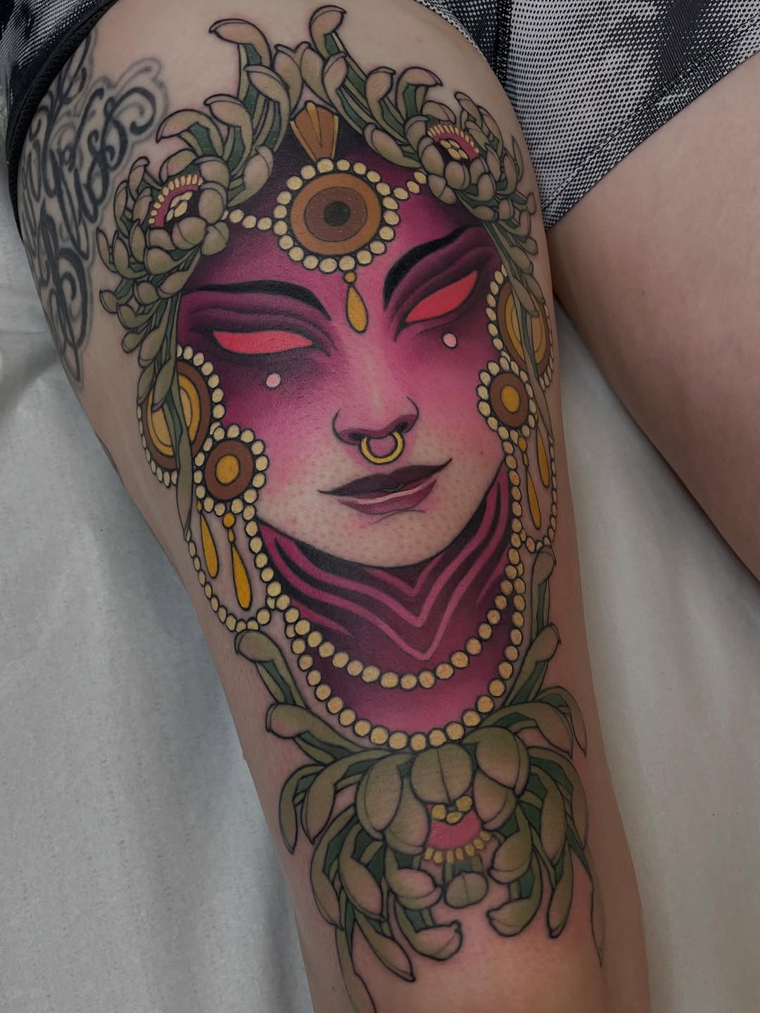 Colorful tattoo on thigh featuring a magenta-faced figure with gold jewelry, ornamental flowers, and decorative elements surrounding the face.