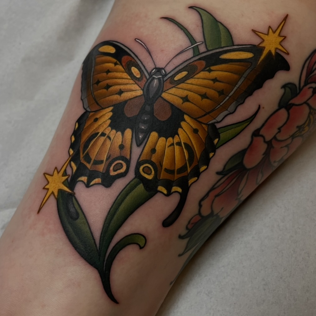 Colorful butterfly tattoo with orange and black wings, green leaves, and gold stars on the inner forearm.