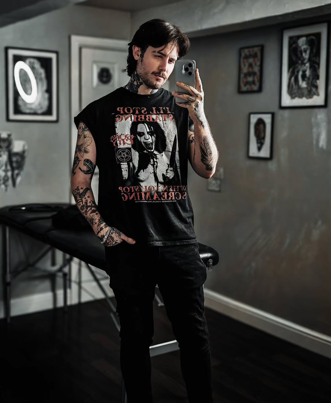 Tattooed man with dark hair holding phone in modern studio, displaying extensive arm and hand tattoos in black ink on pale skin.