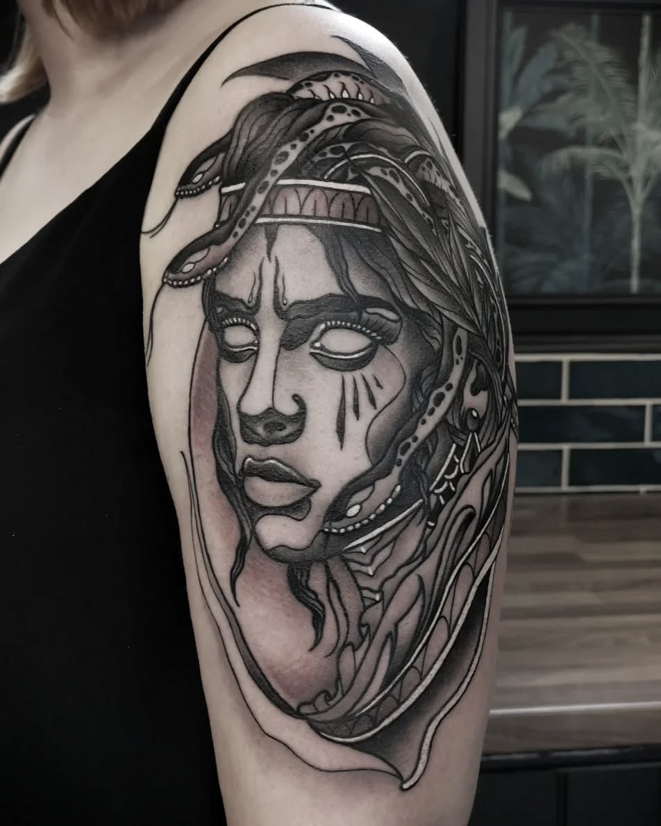 Monochromatic shoulder tattoo depicting a contemplative face with decorative headwear and ornamental details rendered in black ink with shading.