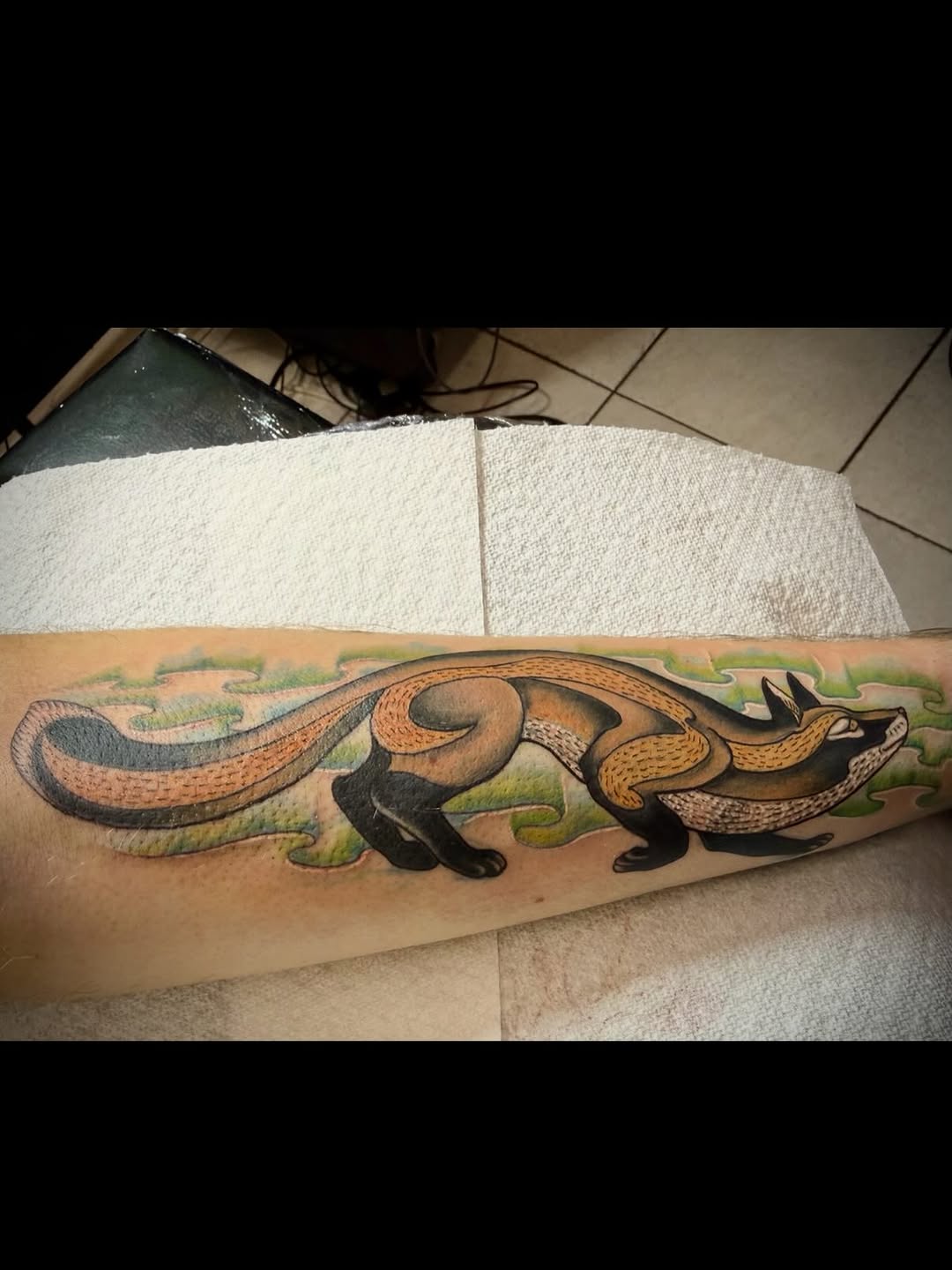 Colorful weasel or ferret tattoo on forearm featuring brown, tan, black, and green tones with grass background detail.