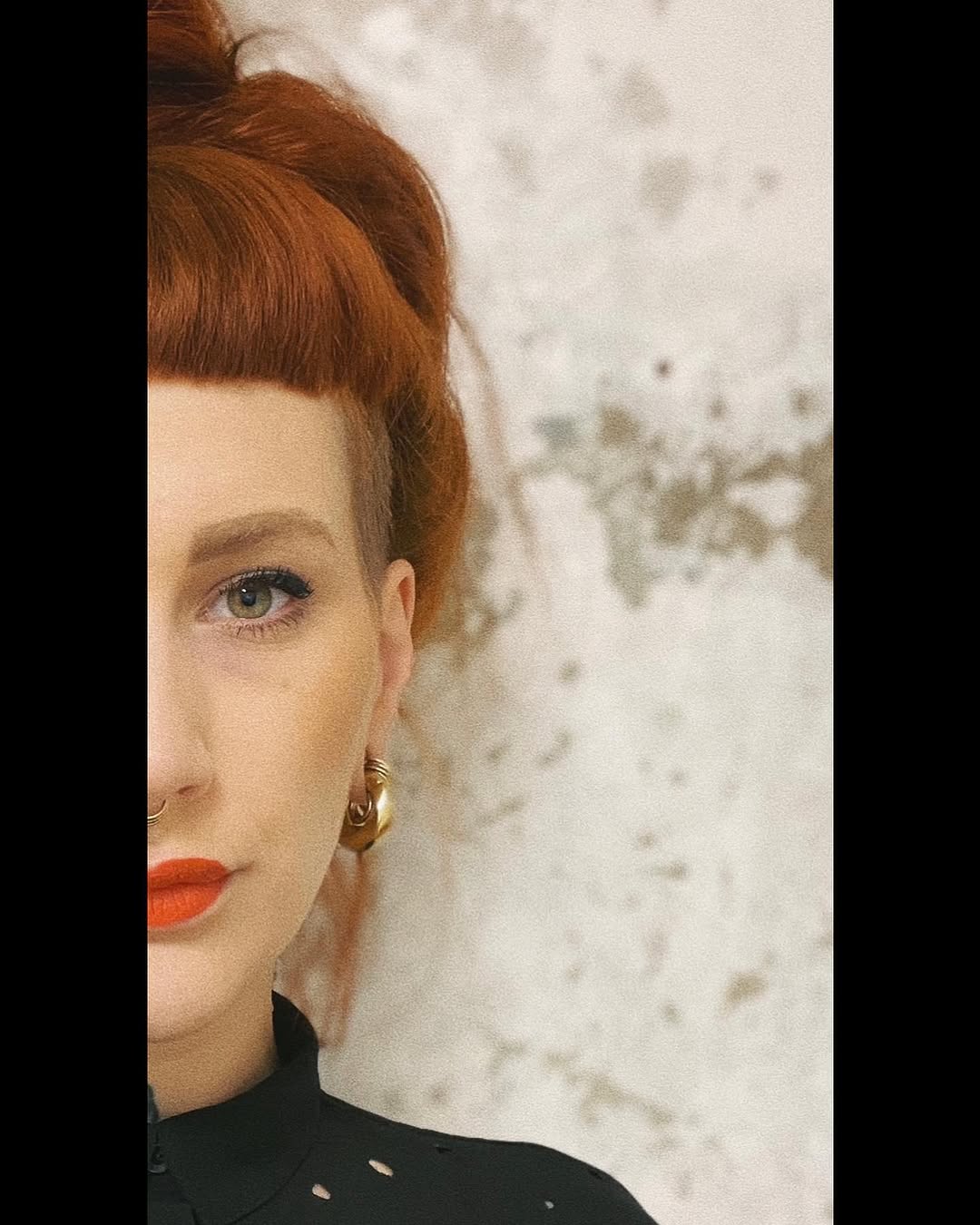 Portrait of a woman with reddish-brown hair styled in a vintage updo, wearing gold hoop earrings and red lipstick against a textured light background.