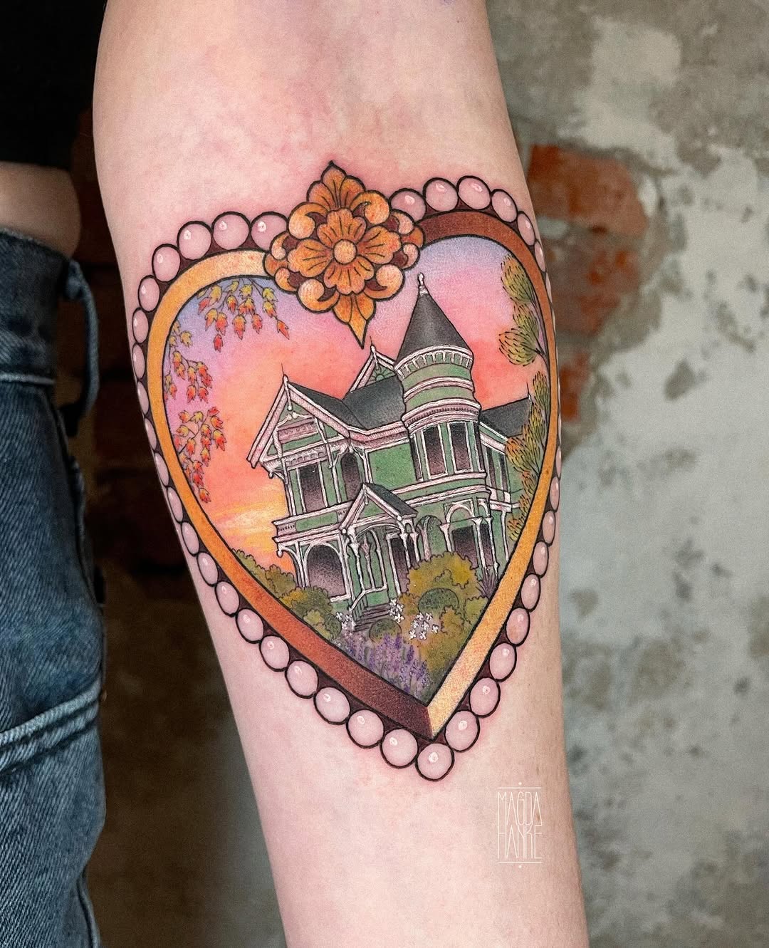 Heart-shaped tattoo with a Victorian mansion, flowering tree, orange flowers, and pearl border on an arm, featuring pink and orange sunset tones.