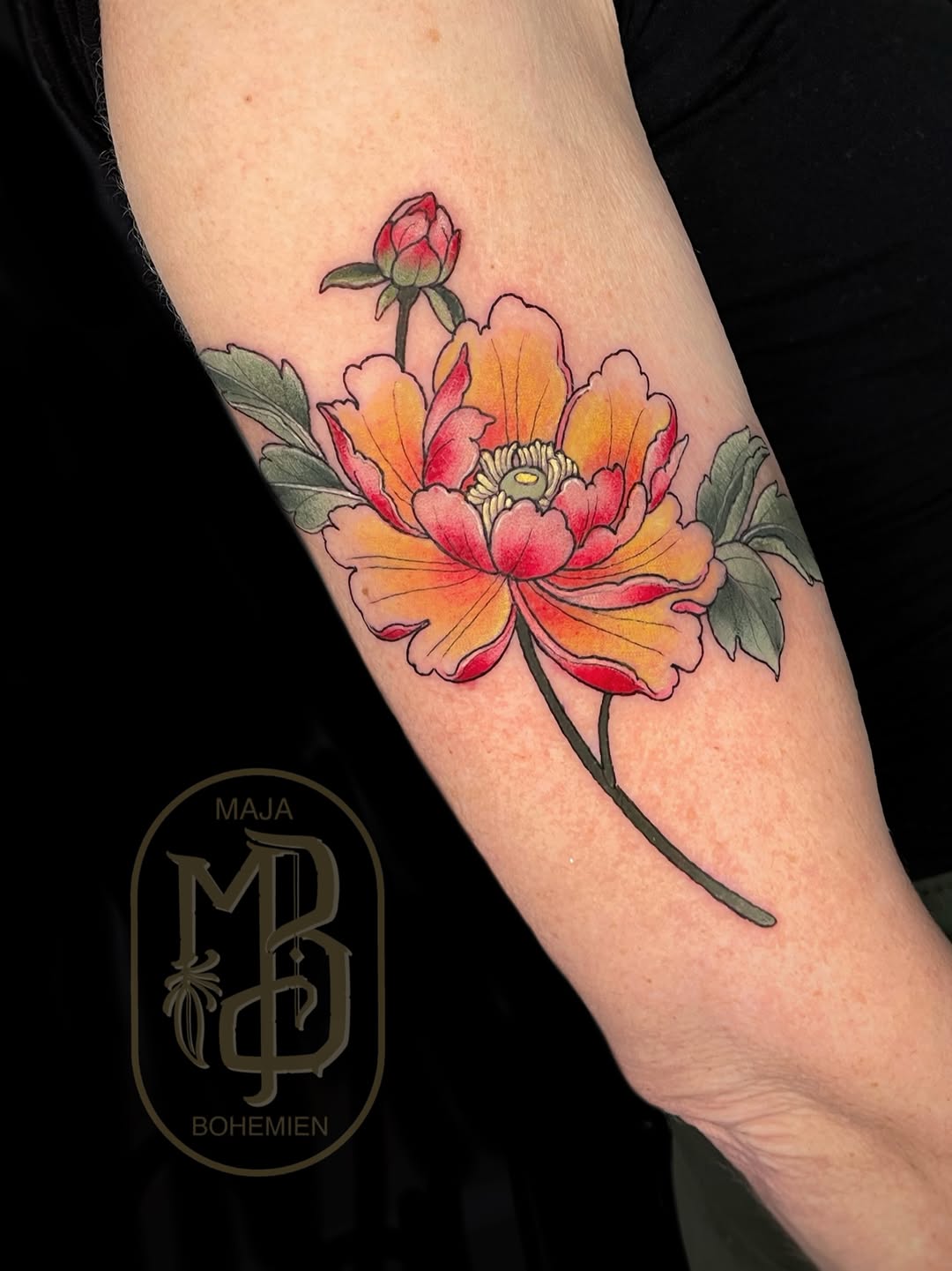 Colorful flower tattoo on the forearm featuring red, orange, and yellow petals with green leaves and a budding stem, signed by Maja Bohemien.