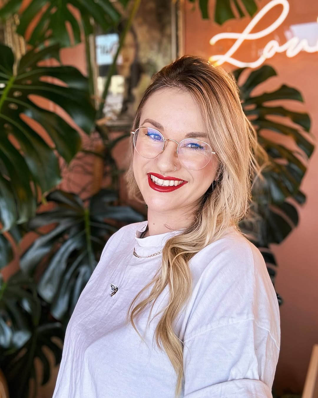 Woman wearing a light purple shirt with a small butterfly tattoo visible on her chest, smiling against an orange wall with potted plants.