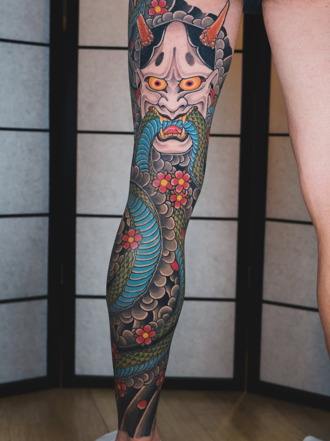 Full leg sleeve tattoo featuring a demon face with orange eyes, serpent body with blue stripes, flowers, and clouds in red, green, and tan colors.