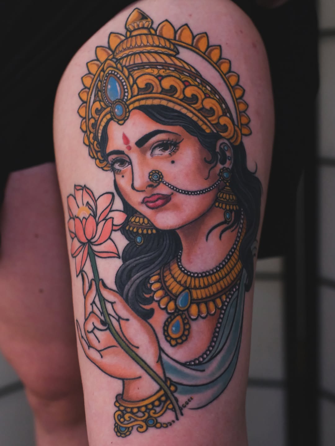 Colorful shoulder tattoo depicting a woman with ornate gold headdress and jewelry, holding a lotus flower, with blue accents and red bindi.