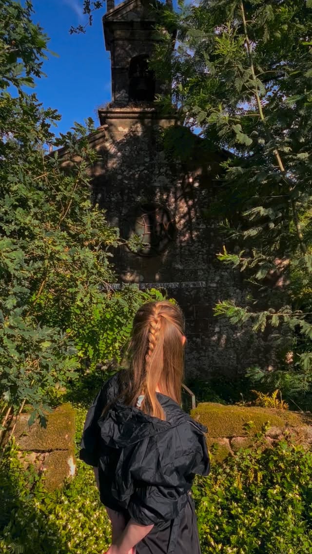 Woman with reddish-brown braided hair wearing a black jacket, viewed from behind facing an ivy-covered stone tower structure against a blue sky.