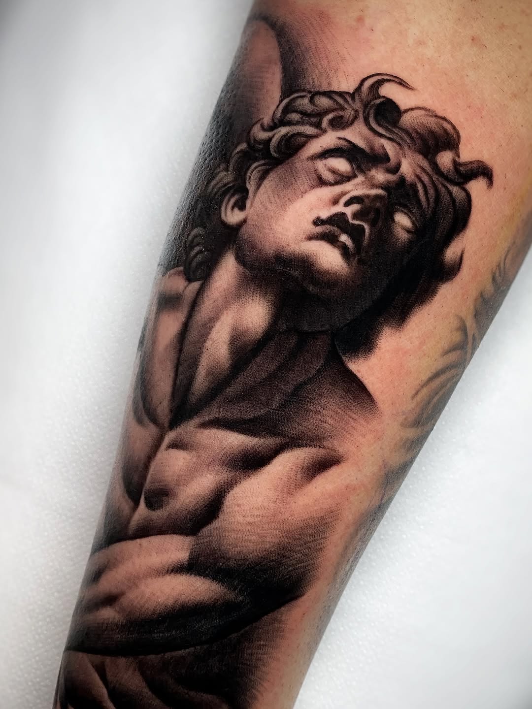 Classical male bust sculpture tattoo on forearm in brown and black ink, depicting curly-haired figure with defined musculature in profile.