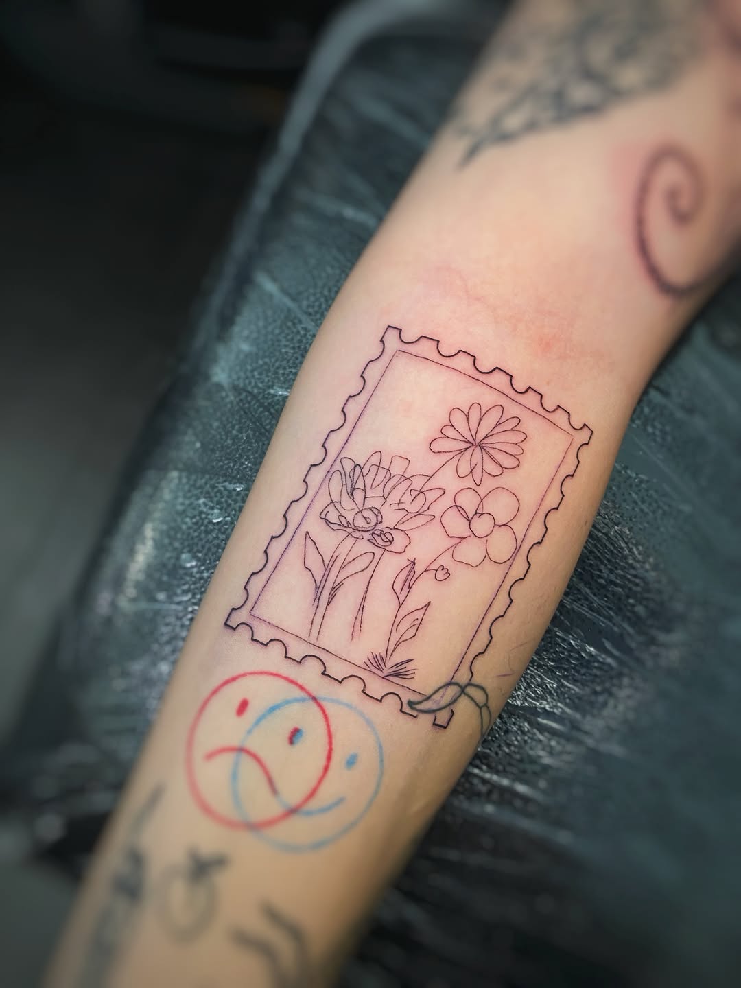 Arm tattoo featuring a stamp-framed bouquet of flowers in pink and purple outlines, with a red and blue smiley face below.