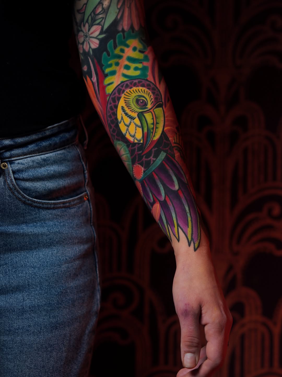 Colorful parrot tattoo on forearm featuring yellow, green, purple, and red hues with floral elements above, displayed on a person wearing denim.