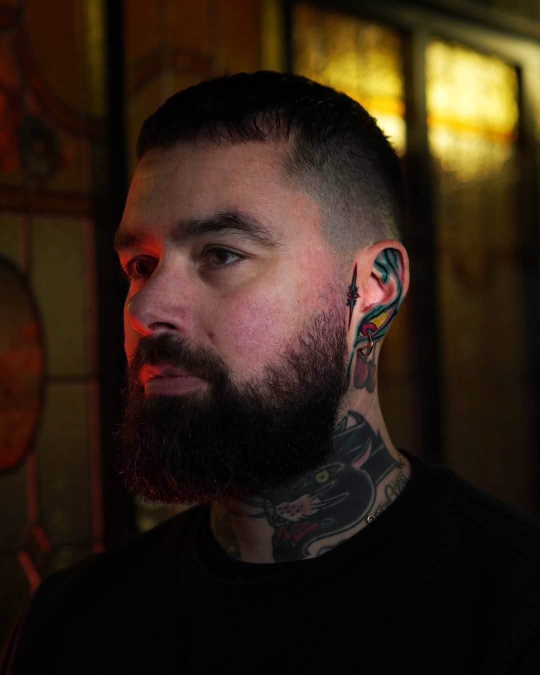 Man with full neck tattoo featuring colorful bird and floral designs, beard, short dark hair, photographed in profile against warm indoor lighting.