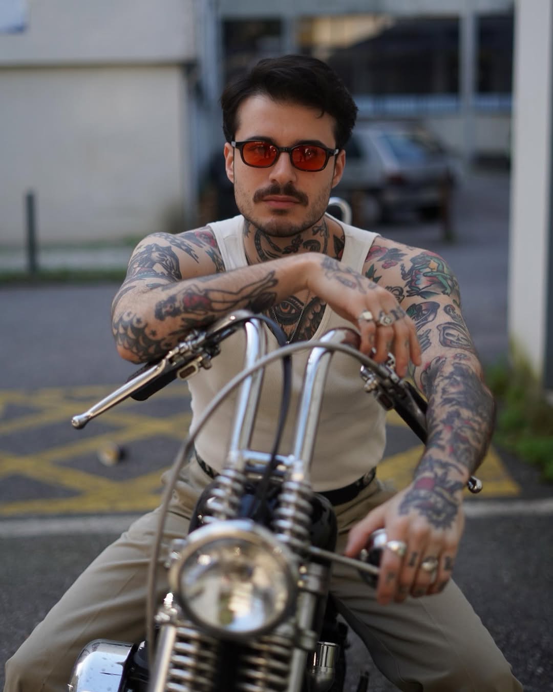 Tattooed man with colorful sleeve tattoos on both arms posing on a motorcycle, wearing sunglasses and a cream-colored shirt.