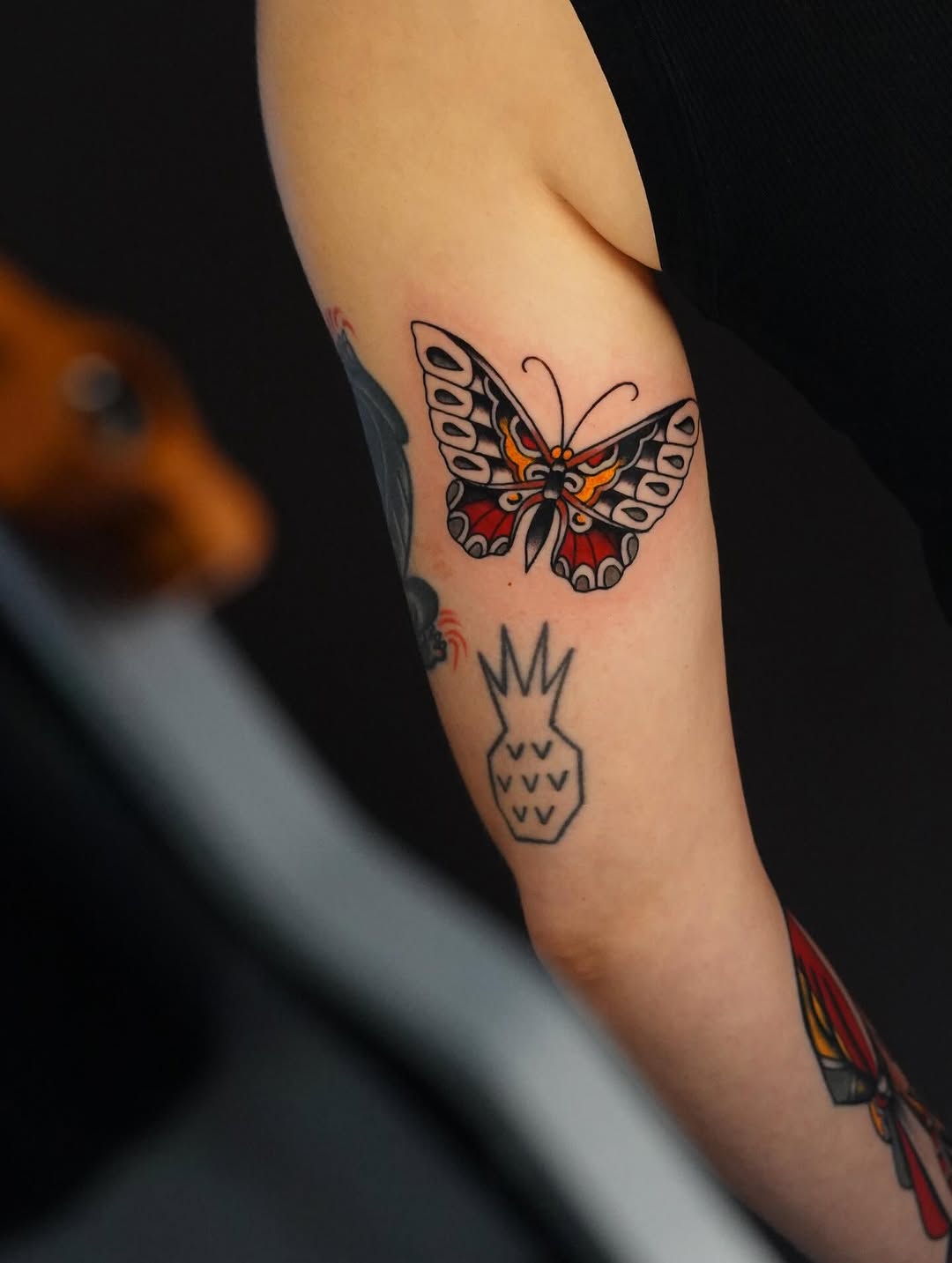Colorful butterfly tattoo with orange, red, and black details on the inner arm, with a small pineapple outline below it.