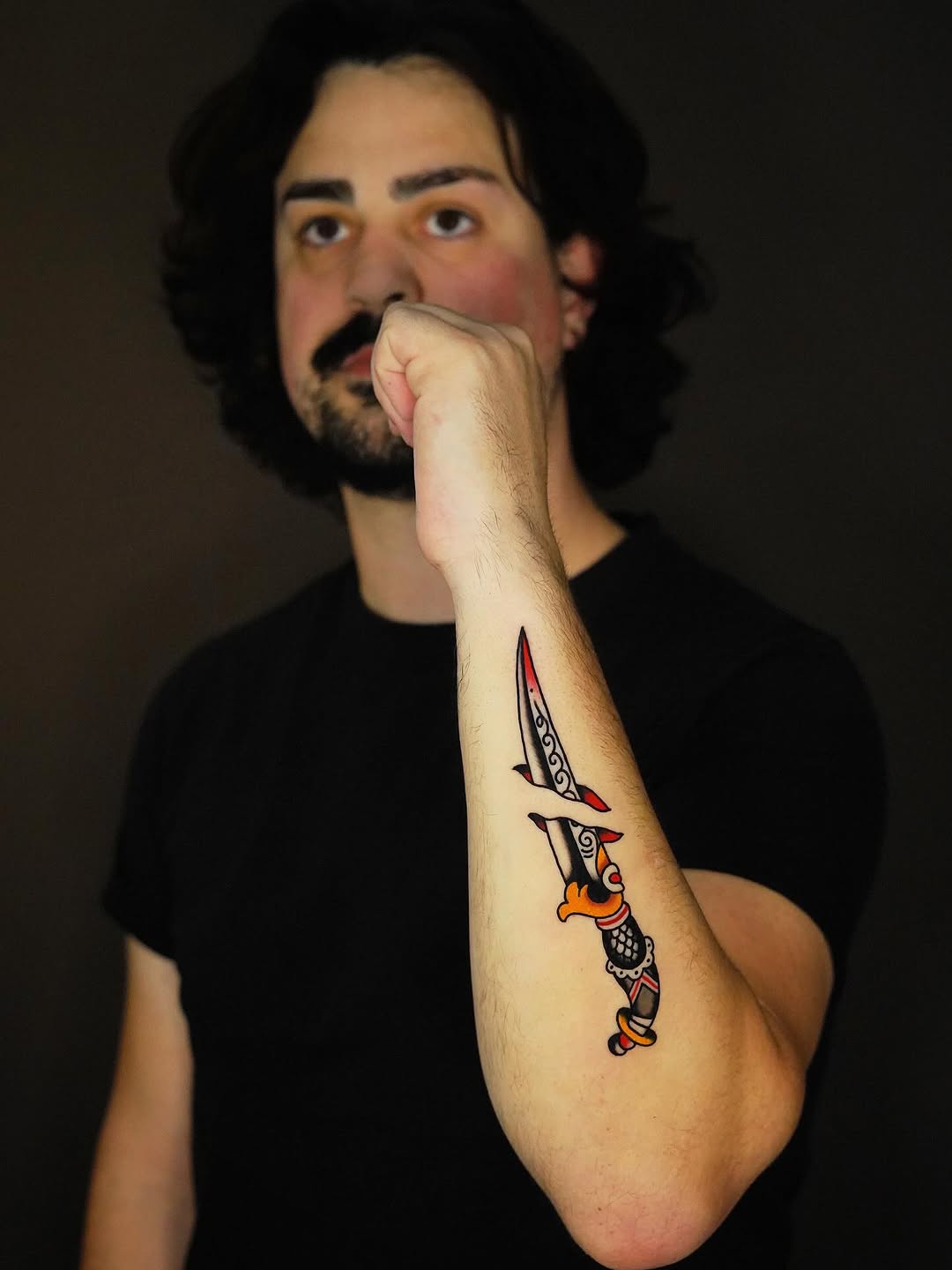 Man displaying a colorful dagger tattoo on his inner forearm featuring red, orange, and black details with decorative bands.