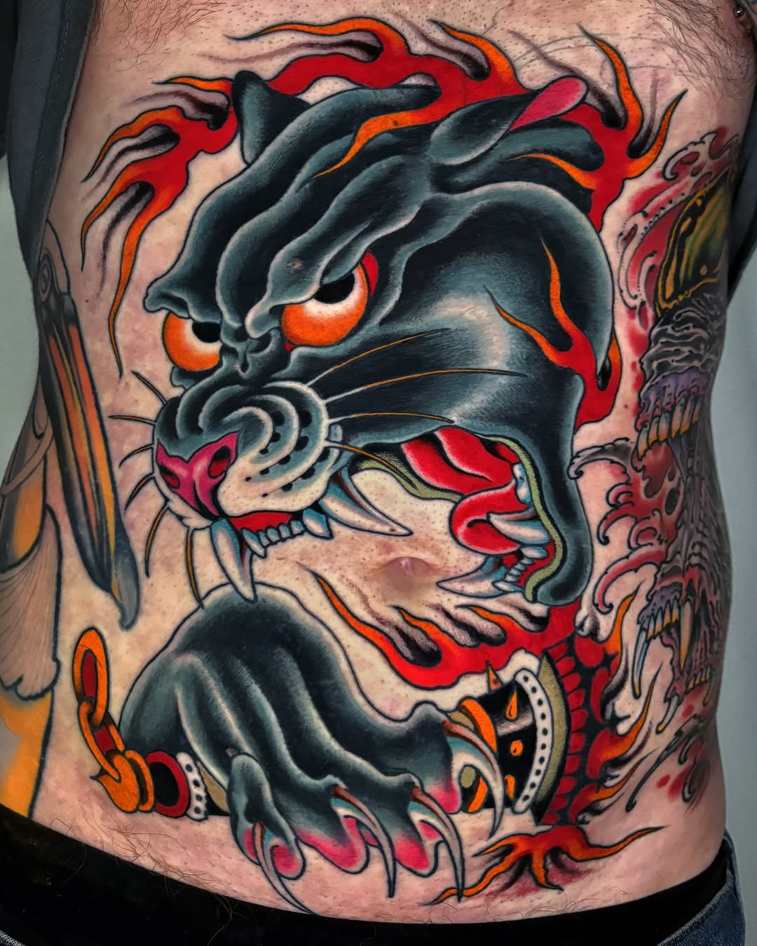 Demonic face tattoo with glowing orange eyes, red flames, sharp teeth, and decorative elements in black, red, orange, and gray on the chest.