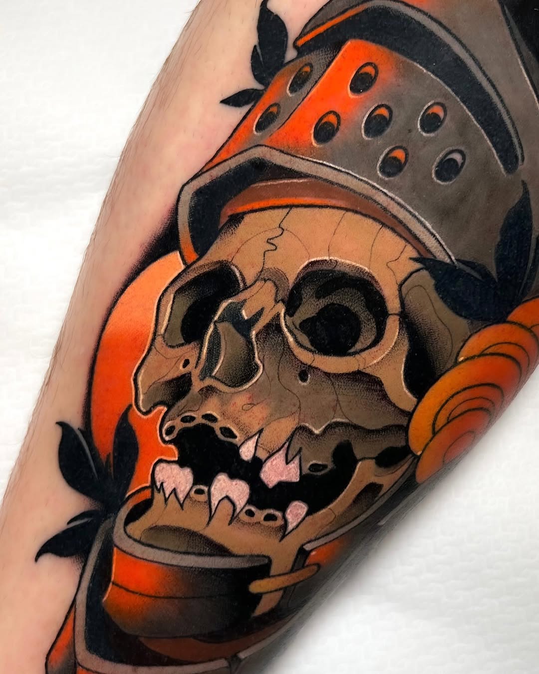 Skull tattoo in orange, brown, and black with a steampunk hat and floral elements on the upper arm.