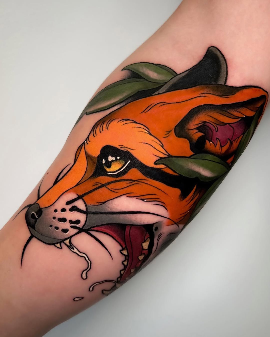 Orange fox head with green leaves, open mouth showing pink interior, yellow eye, and black whiskers tattooed on an arm.